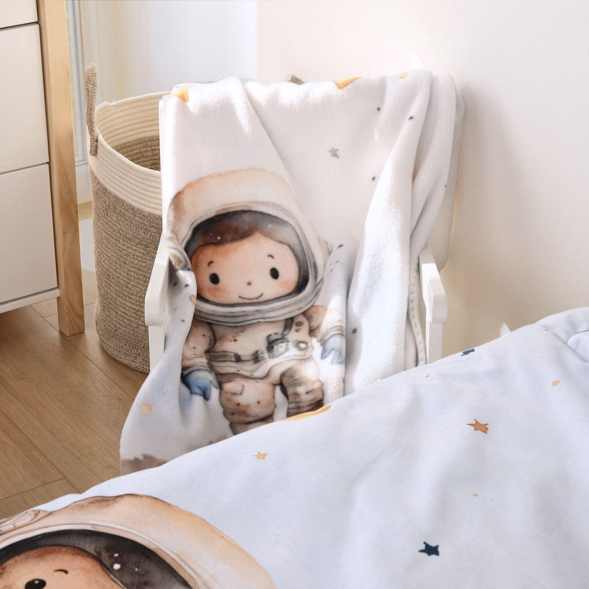 babybest Kuscheldecke ‘Kleiner Astronaut’ über einem Stuhl neben einem geflochtenen Korb