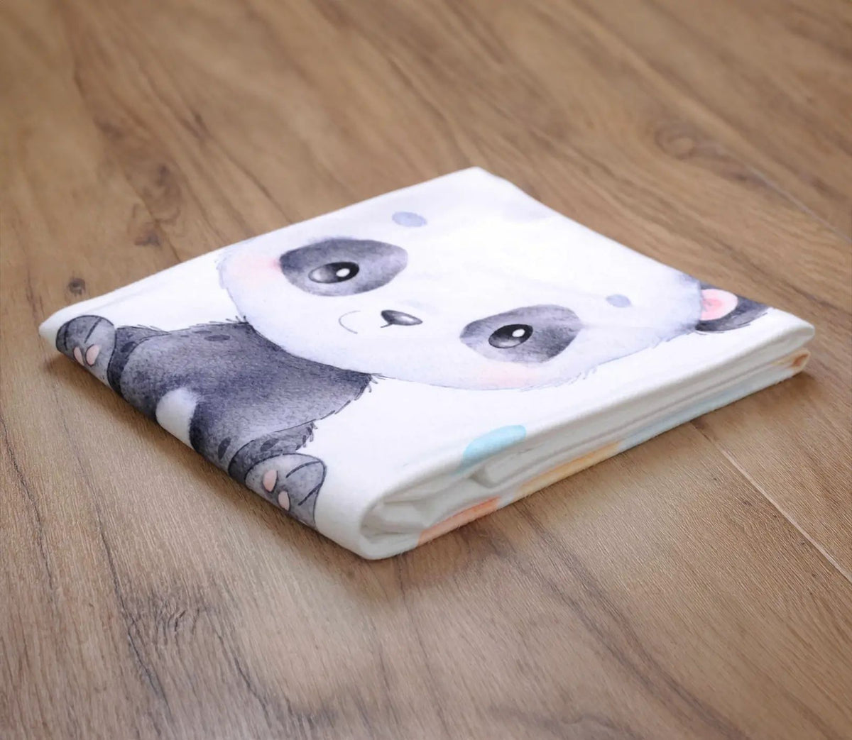 Zusammengelegte Kuscheldecke ‘Regenbogen Panda’ auf einem Holzboden