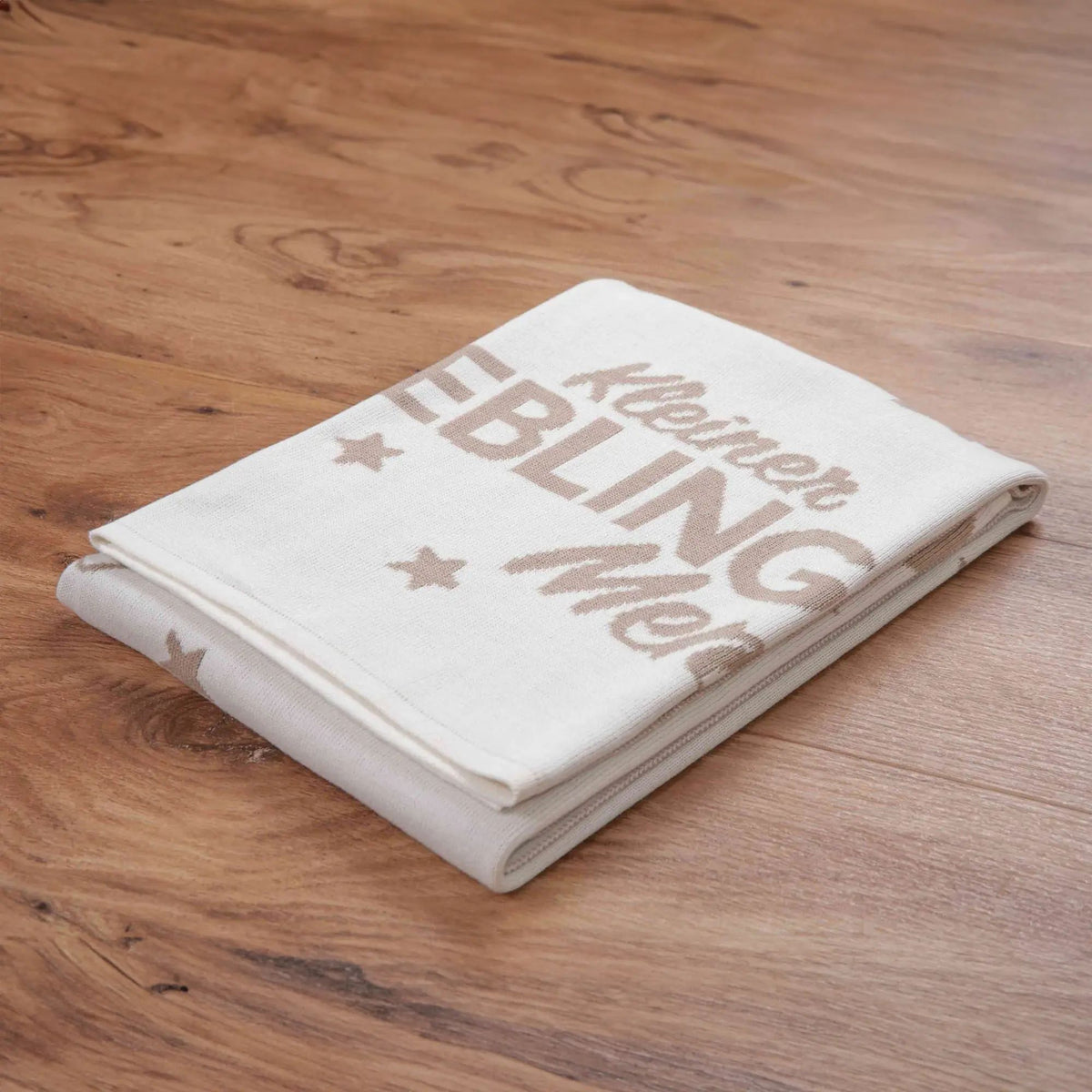 Zusammengelegte Strickdecke ‘Kleiner Lieblingsmensch’ mit Schriftzug auf einem Holzboden