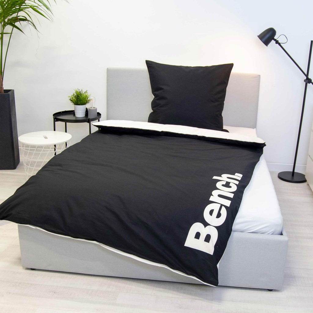 Bench Bettwäsche ‘Schwarz/Weiß’ auf einem grauen Polsterbett, schwarze Seite mit weißem Logo