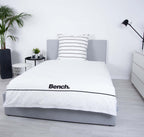 Bench Bettwäsche ‘Weiß/Gestreift’ auf einem grauen Polsterbett, Logo-Seite