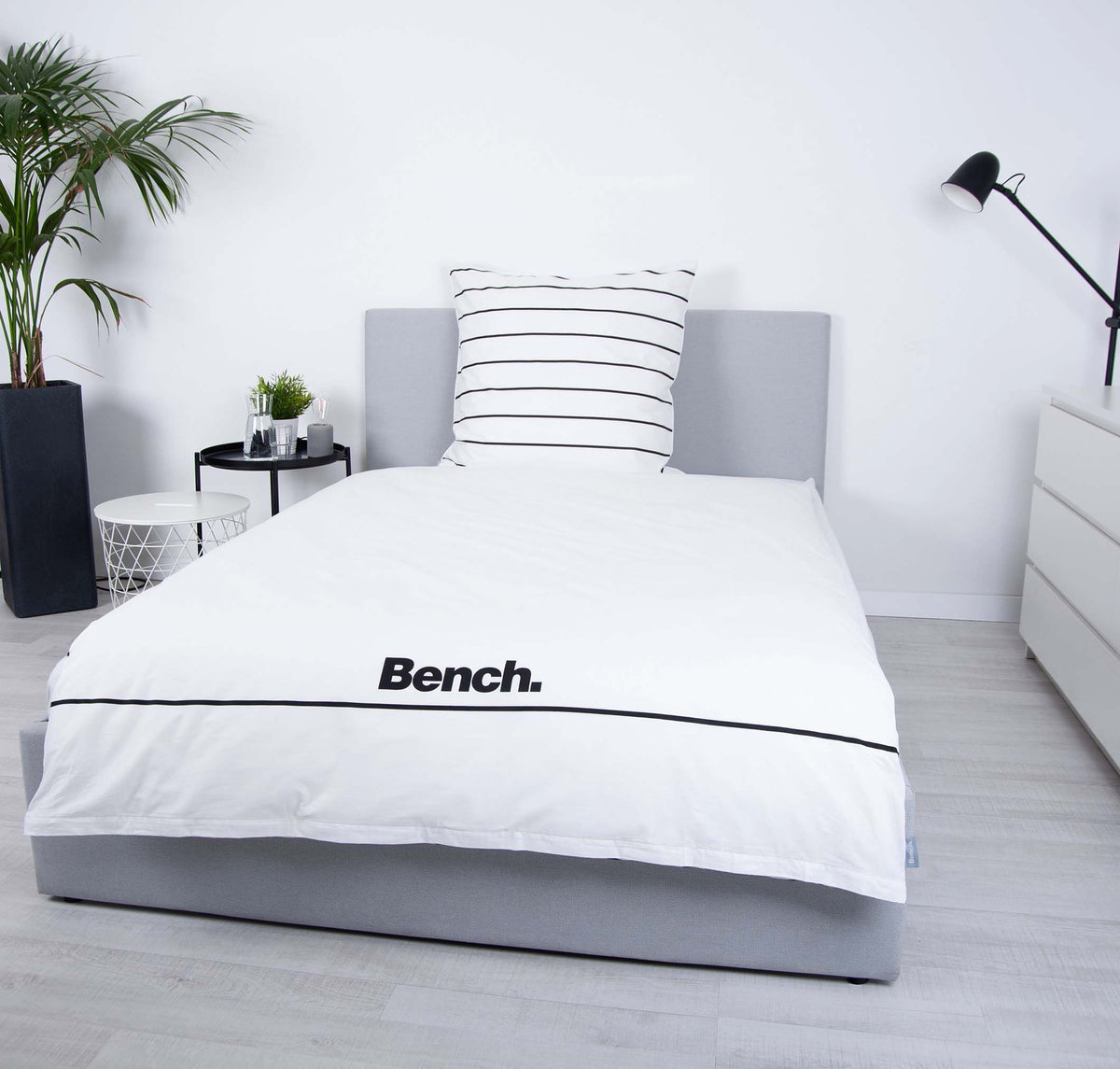 Bench Bettwäsche ‘Weiß/Gestreift’ auf einem grauen Polsterbett, Logo-Seite