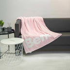 Bench Kuscheldecke in Hellrosa mit Logo auf einem dunkelgrauen Sofa