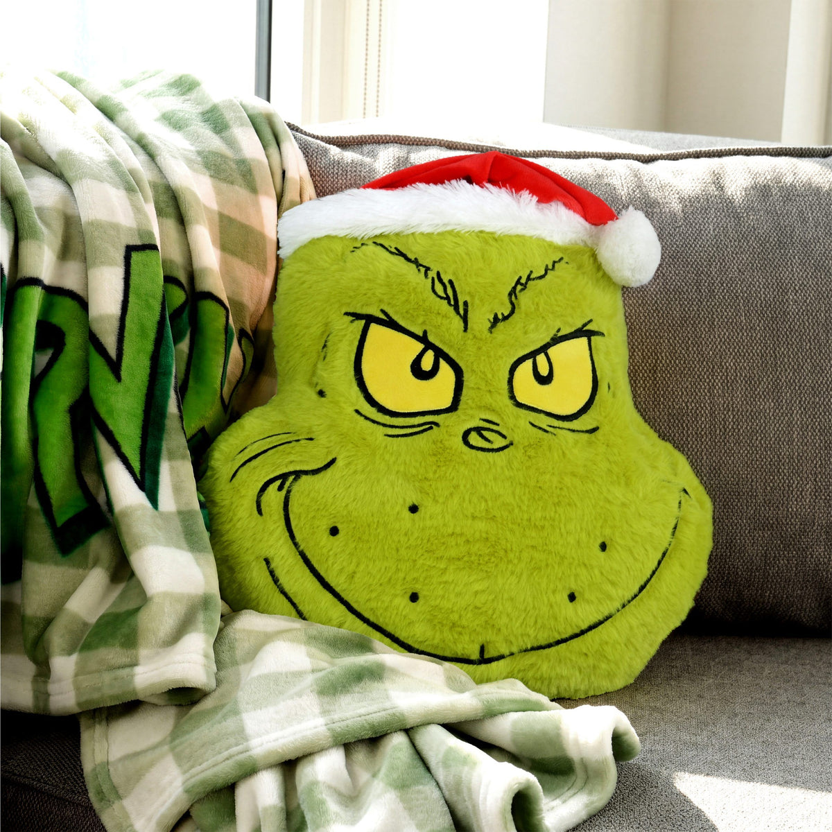 Grinch Formkissen ‘Merry Grinchmas’ auf dem Sofa neben grün karierter Grinch-Kuscheldecke