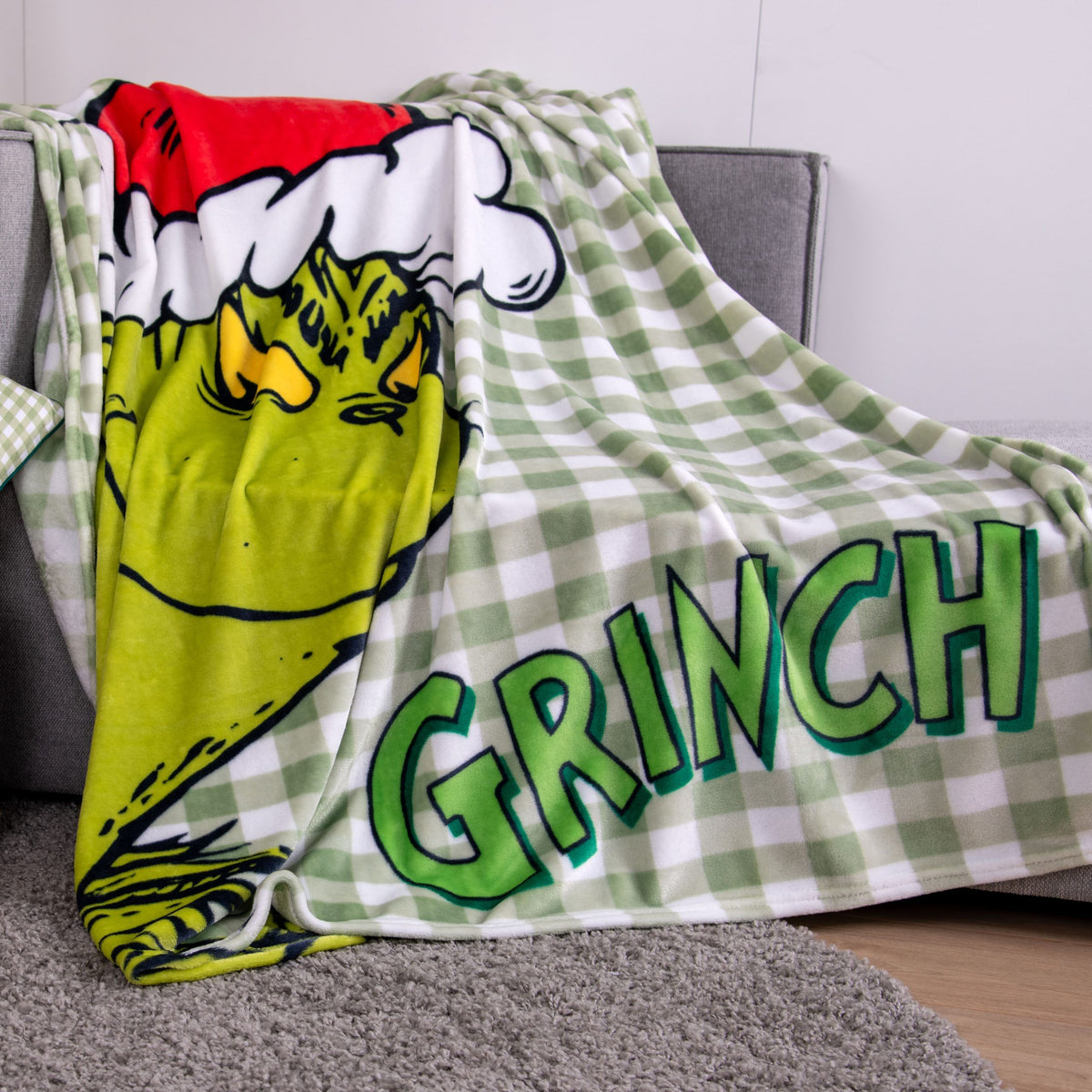 Grinch Kuscheldecke ‘Merry Grinchmas’ über ein graues Sofa gelegt