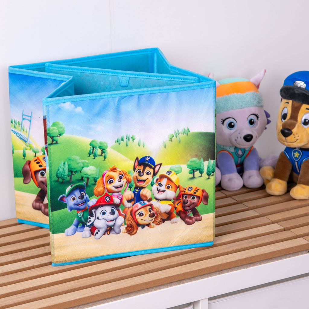 Halb zusammengefaltete Paw Patrol Aufbewahrungsbox mit Marshall, Skye, Chase, Rubble und weiteren Fellfreunden vor grünen Hügeln, daneben Plüschfiguren