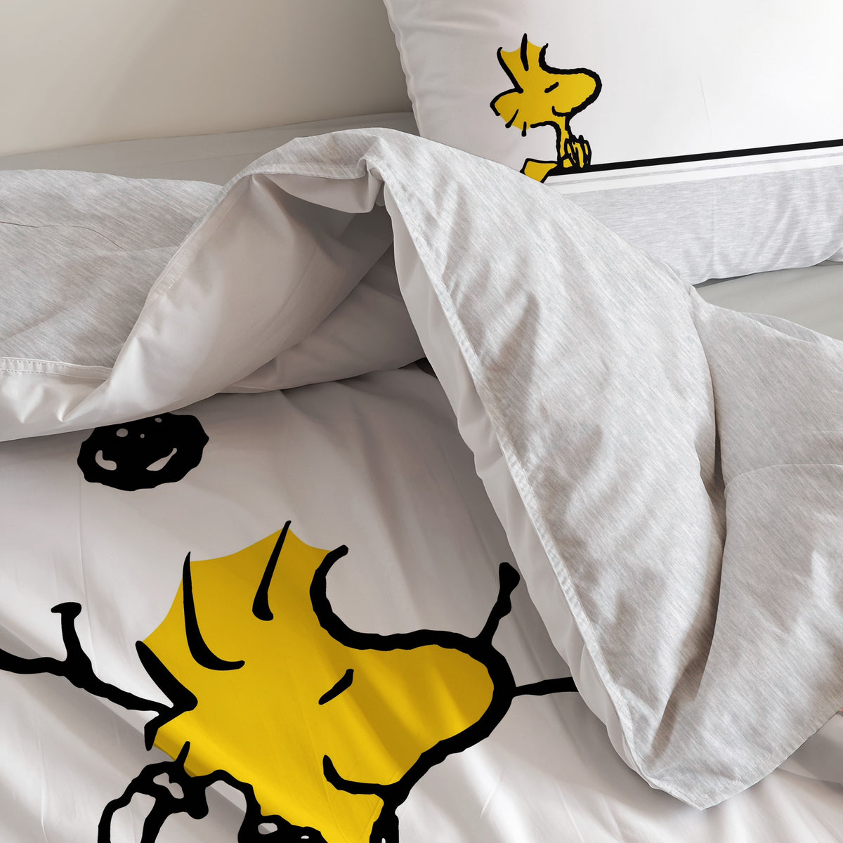 Snoopy Bettwäsche ‘Classic Grey’ auf dem Bett: Woodstock in Snoopys Armen auf dem Bettbezug, dahinter der Kissenbezug mit Woodstock auf dem Streifen
