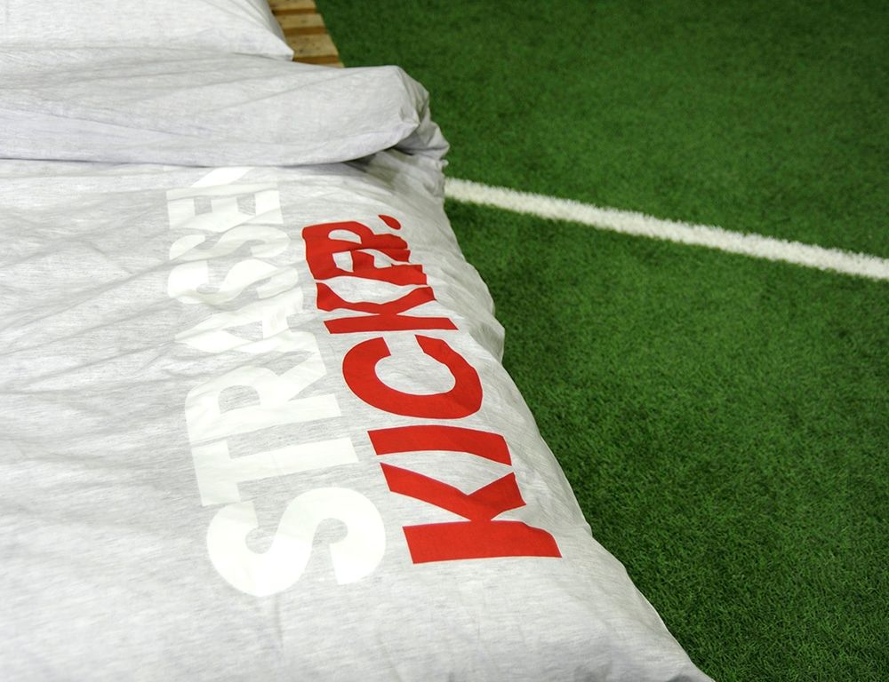 STRASSENKICKER-Logo in Weiß und Rot auf dem hellgrauen Bettbezug, auf Rasen liegend