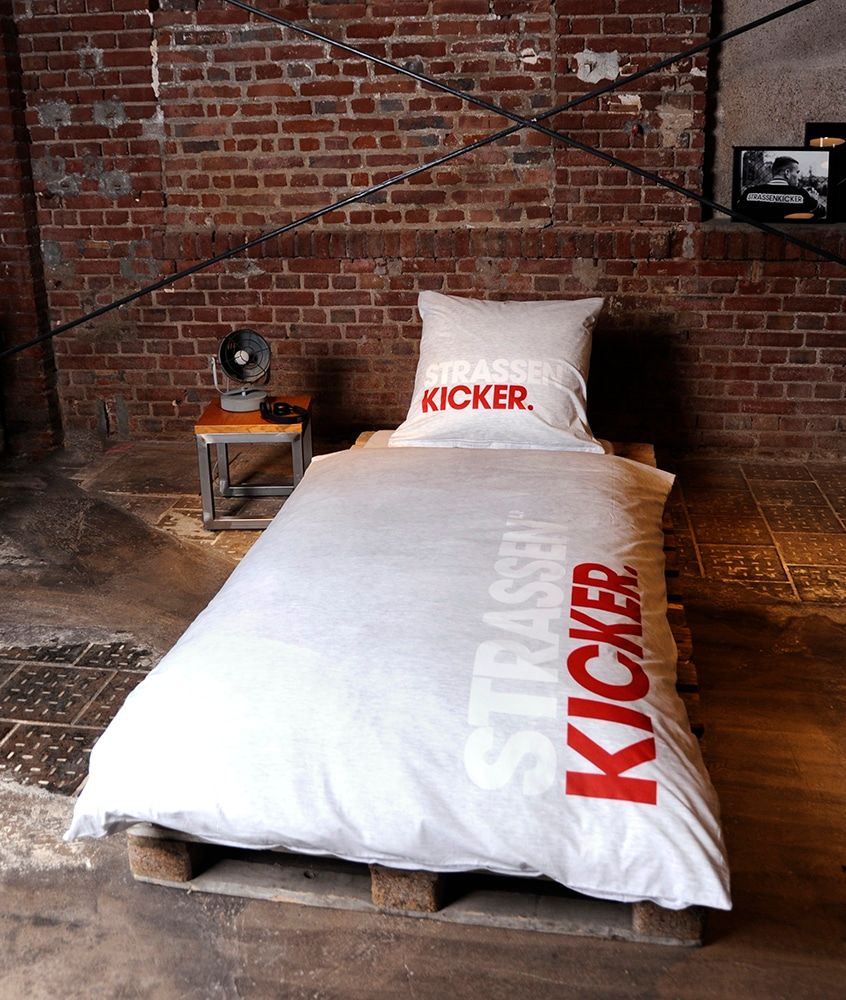 STRASSENKICKER Bettwäsche ‘Hellgrau’ auf einem Bett vor Backsteinwand