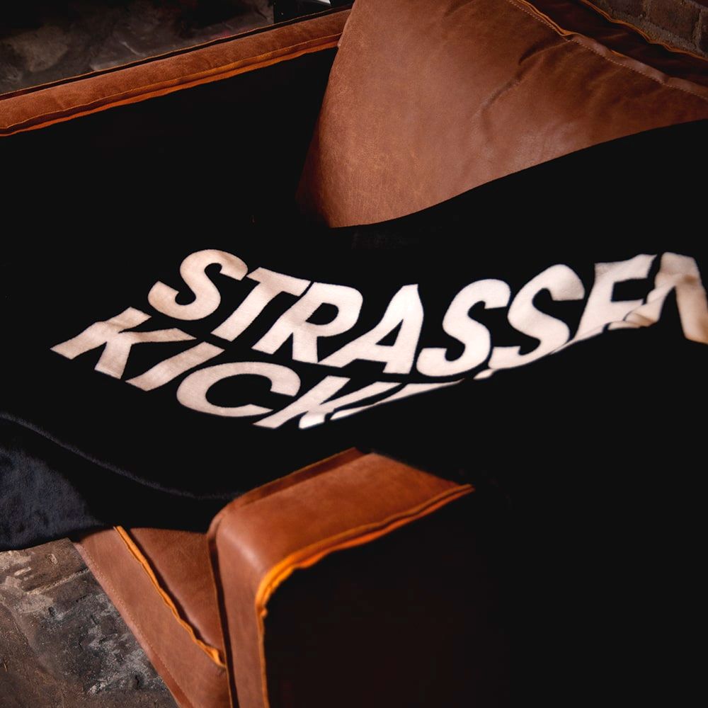 STRASSENKICKER Kuscheldecke auf einem braunen Ledersofa