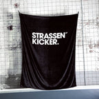 STRASSENKICKER Kuscheldecke an einem Netz vor einer weißen Backsteinwand aufgehängt