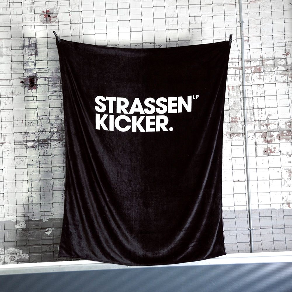 STRASSENKICKER Kuscheldecke an einem Netz vor einer weißen Backsteinwand aufgehängt