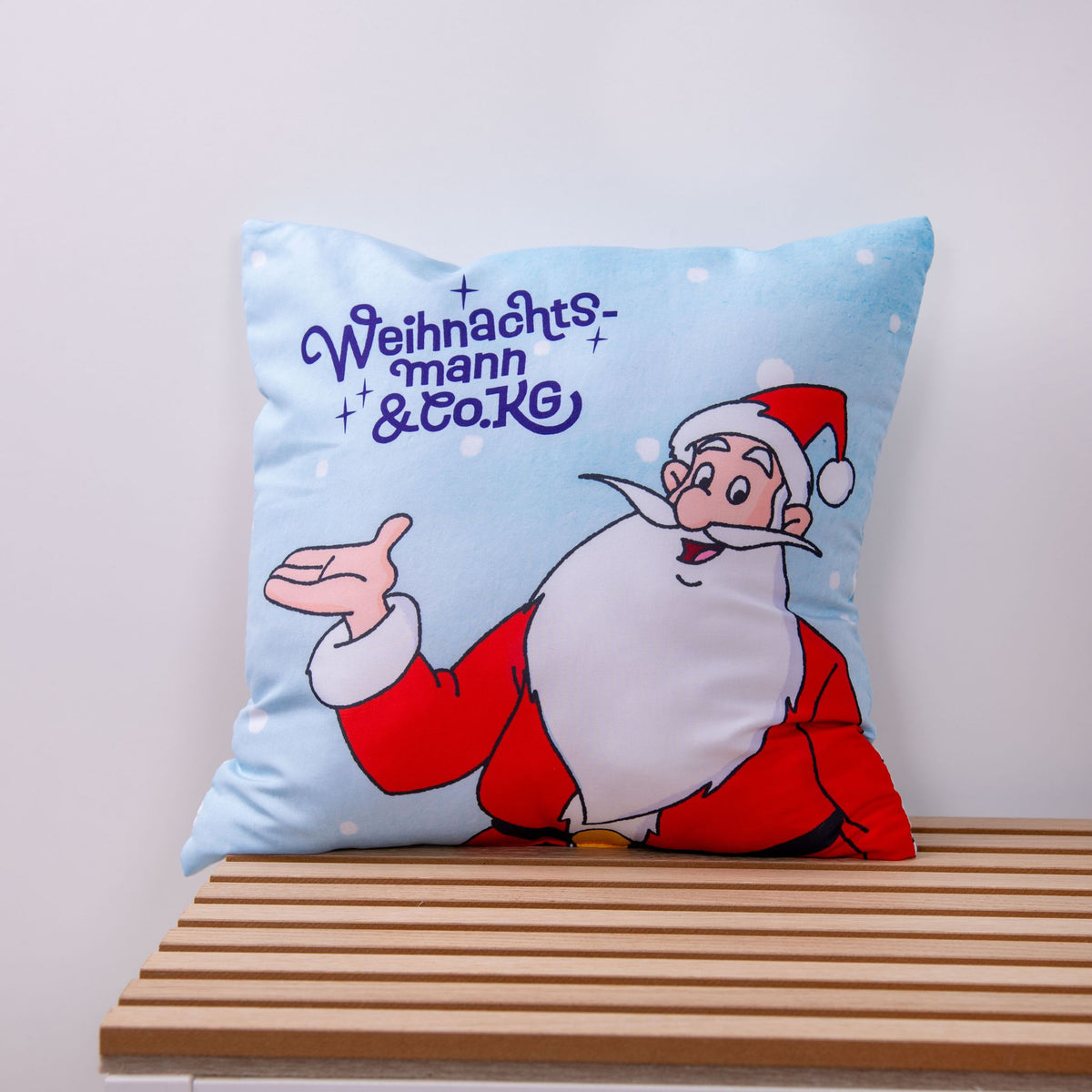 Hellblaues Kissen ‘Nordpol-Helden’: der Weihnachtsmann mit ausgestreckter Hand neben dem lila Schriftzug Weihnachtsmann & Co. KG