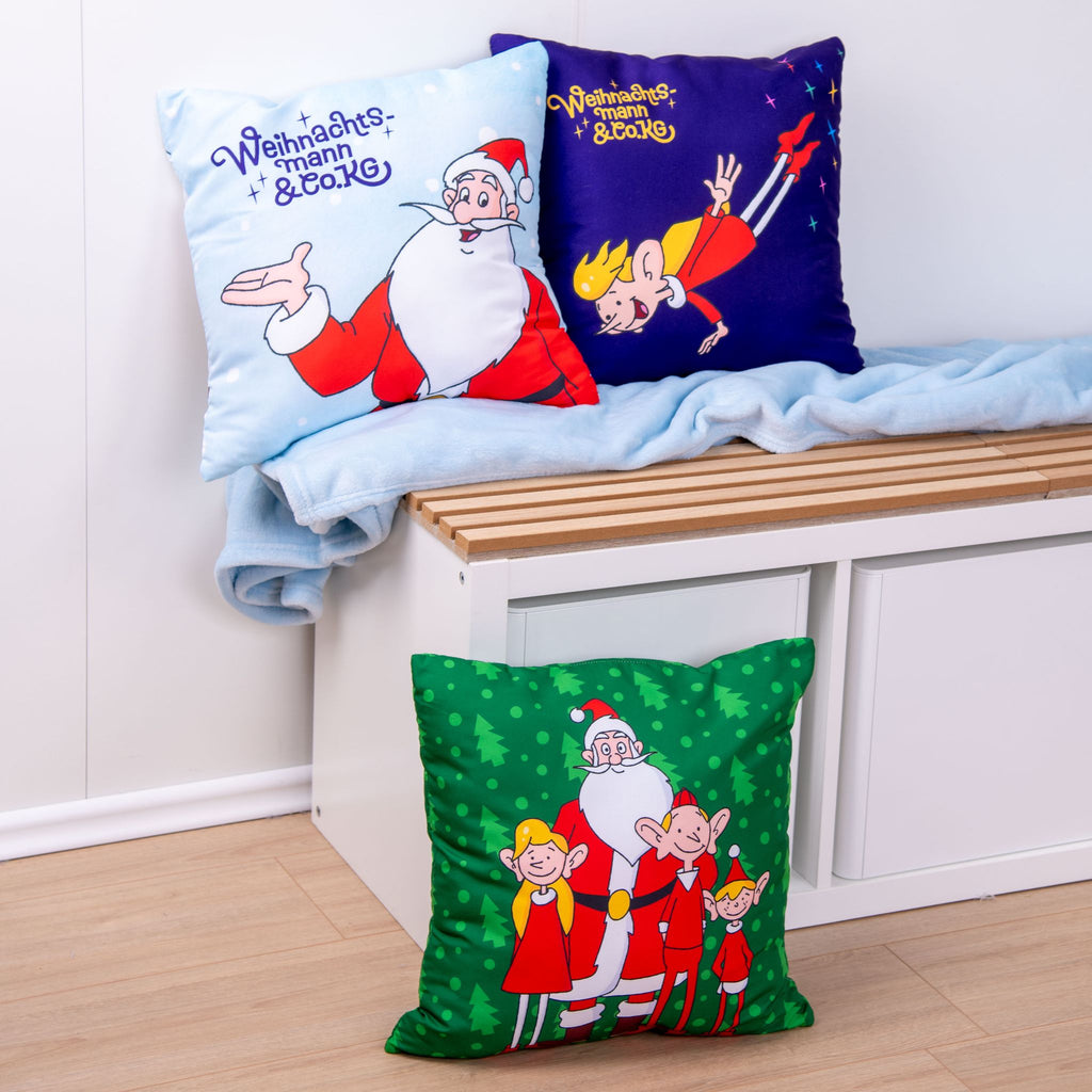 Die Wendeseiten der drei Kissen ‘Nordpol-Helden’ auf der Bank: der Weihnachtsmann mit Schriftzug auf Hellblau, die fliegende Elfe auf Lila und der Weihnachtsmann mit drei Elfen auf Grün