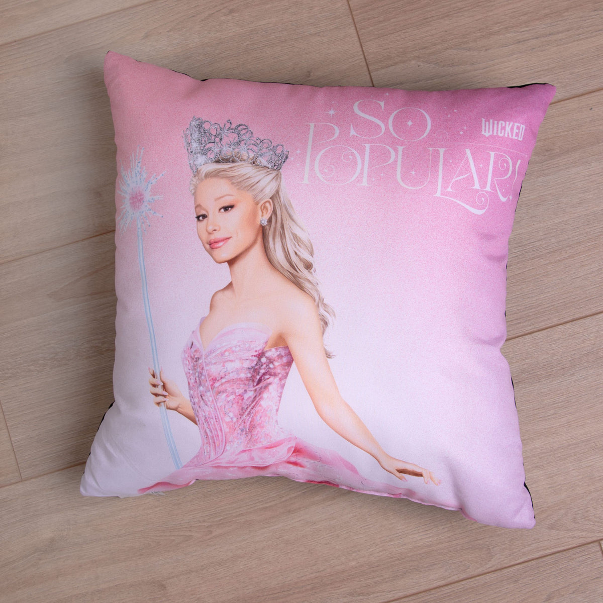 Kissen mit Glinda und dem Schriftzug „So Popular!“ auf einem Holzboden
