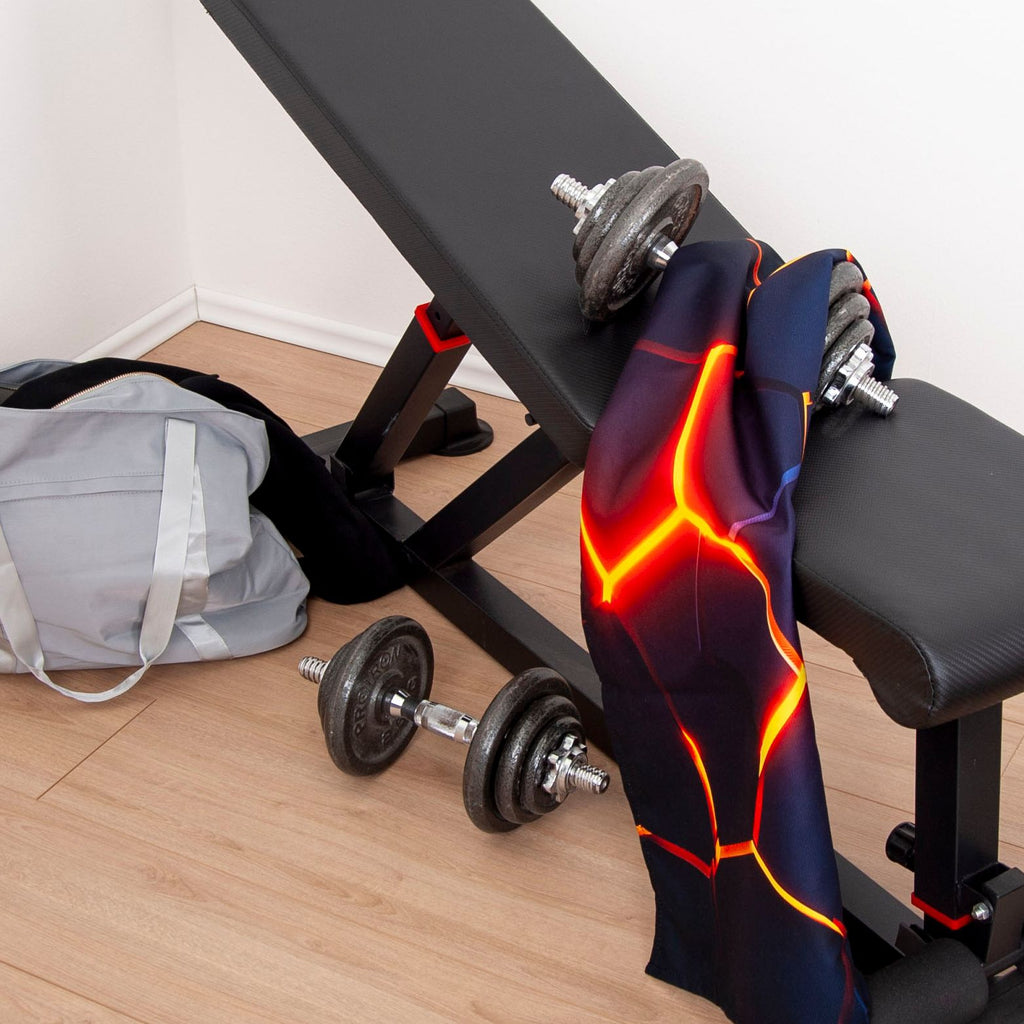 Fitnesstuch ‘Lava 3D Effekt’ über einer Kurzhantel auf der Hantelbank, daneben eine zweite Hantel und eine Sporttasche